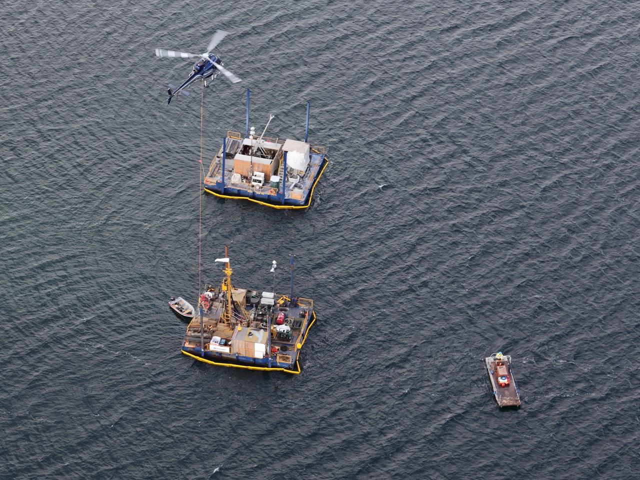aerial view helicopter lowering diamond drilling equipment to large water barge below with two other water barges floating close by